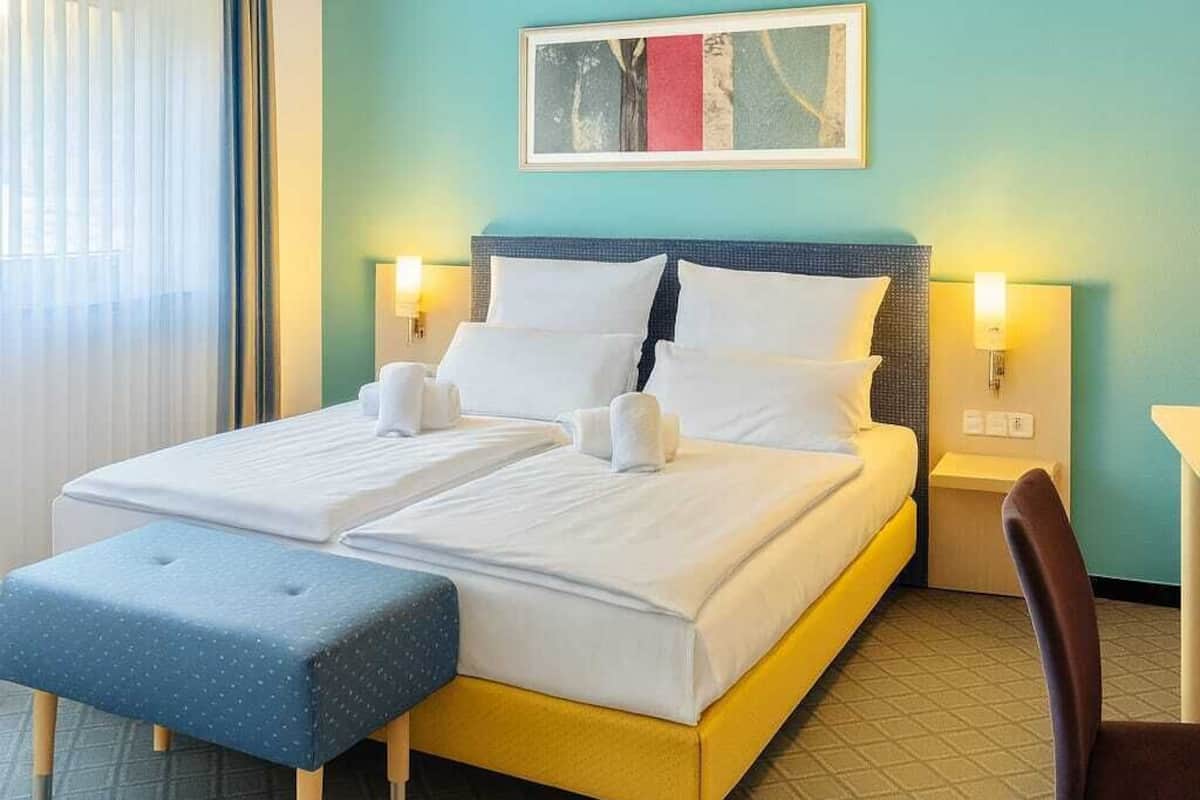 A hotel room with a double bed, a small table, a chair, and a window with curtains.