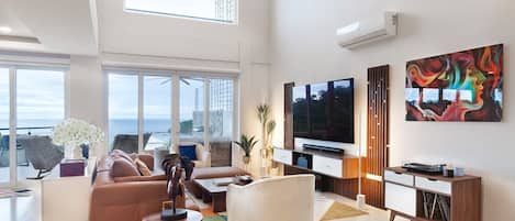 A modern living room with a large TV, comfortable seating, and a view of the ocean.