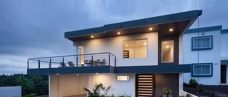 A modern two-story house with a balcony, large windows, and a well-lit entrance.