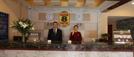 Un comptoir d’accueil d’hôtel avec un revêtement en marbre, deux réceptionnistes, une horloge et une enseigne indiquant le nom de l’hôtel Super 8.