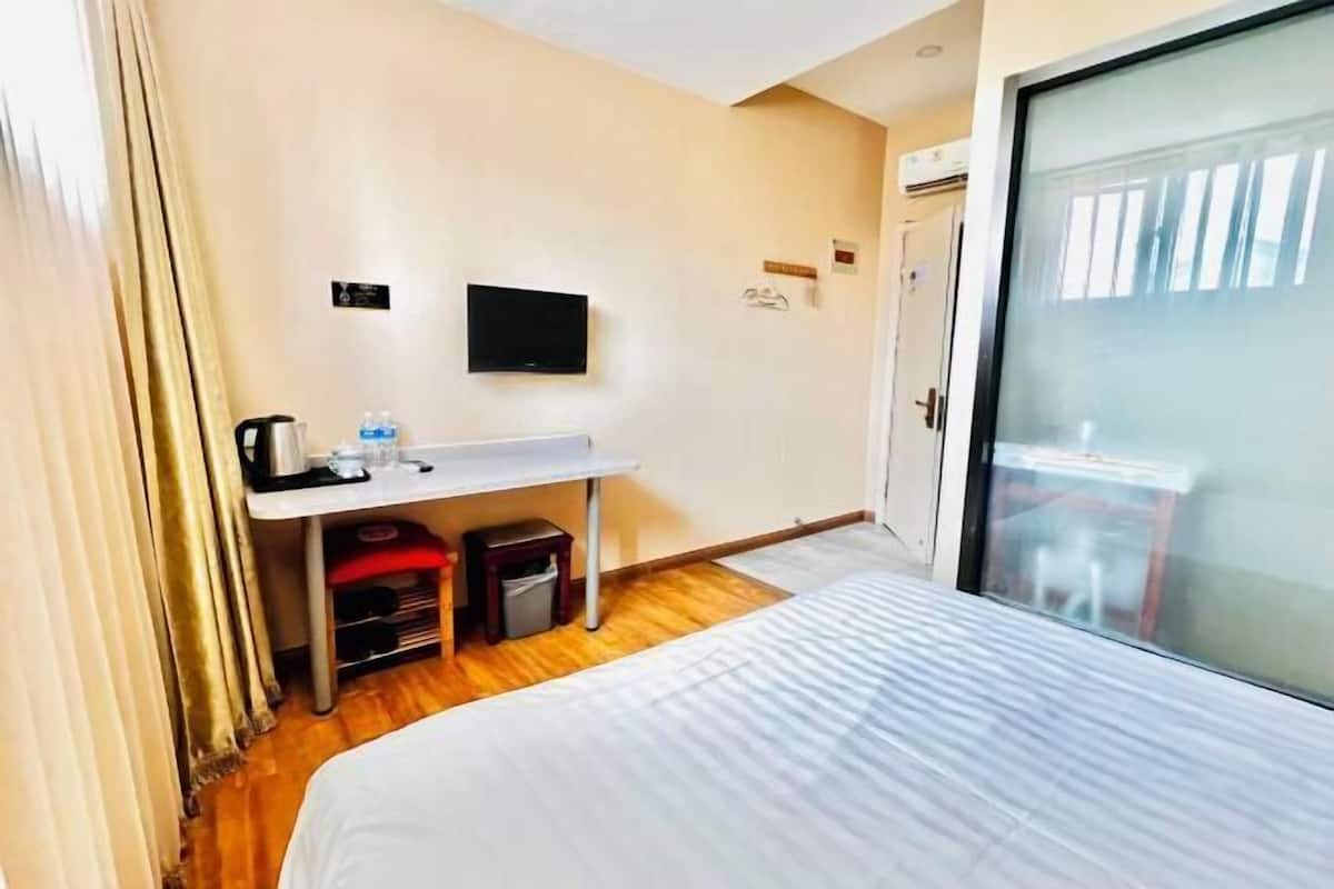A hotel room with a bed, a desk with a flat-screen TV, a small trash bin, and a window with a view.