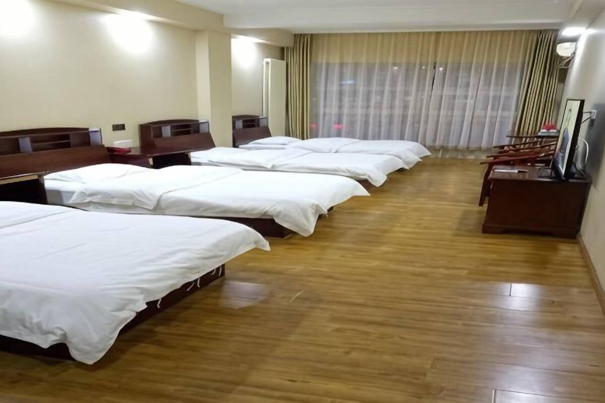 A hotel room with multiple beds, wooden flooring, a TV, and a large window with curtains.