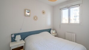 A bedroom with a bed, bedside table, lamp, wall decor, and a window with a view of buildings. - Apartment for 6, ocean view, Mimizan Beach, parking (Mimizan-Mimizan Plage)