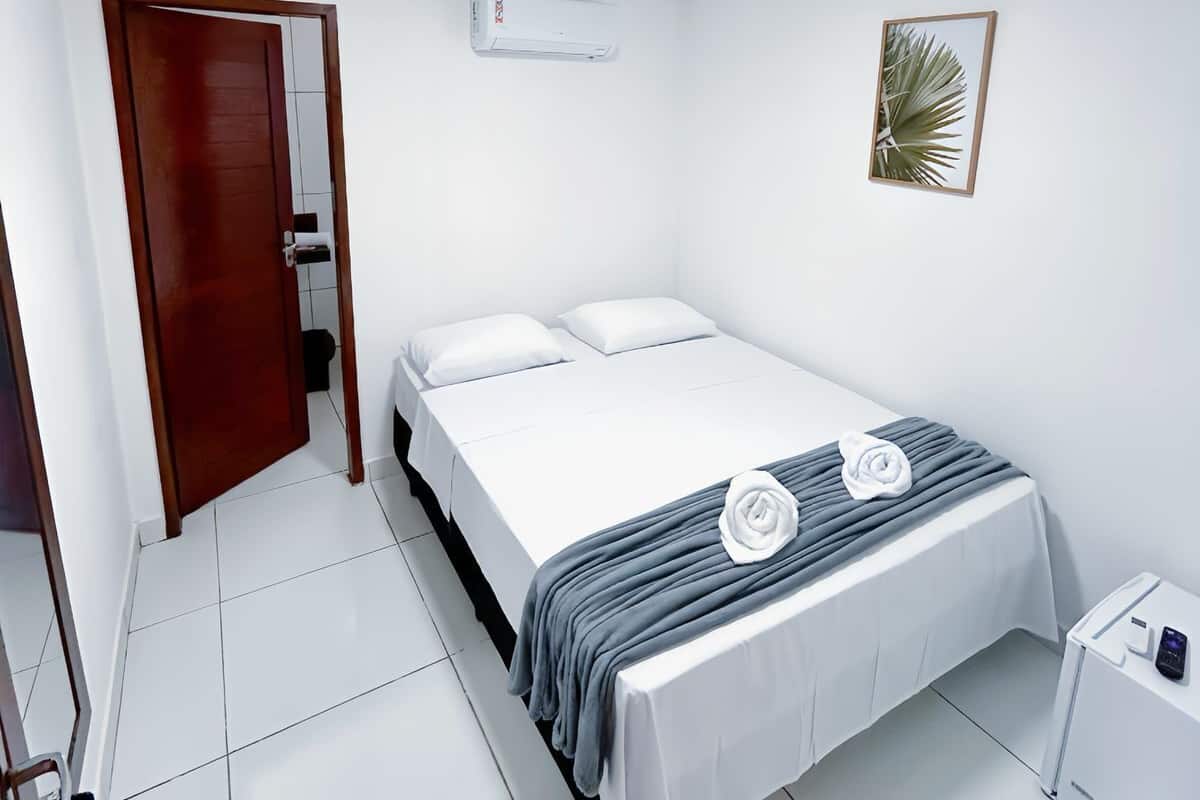 A clean, white room with a bed, a door, an air conditioning unit, and a small table with a phone.