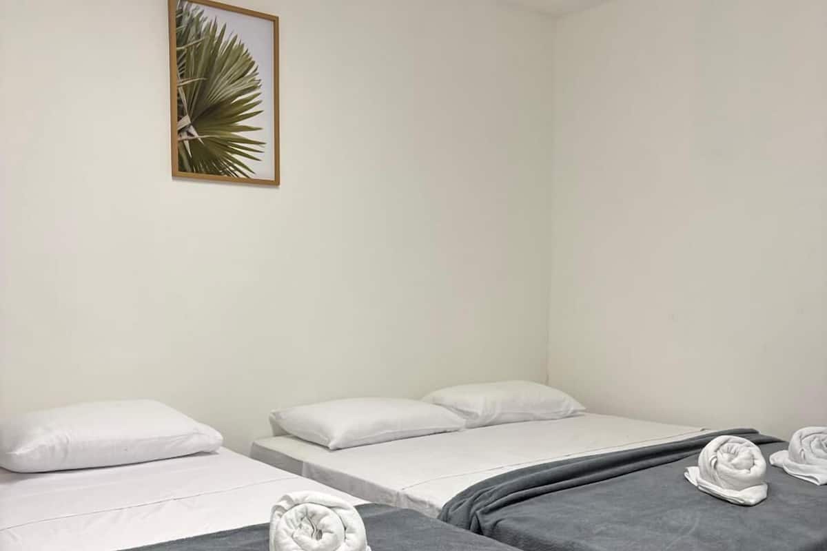 A hotel room with two beds, each with a grey blanket and a rolled towel. There is a framed picture of a palm leaf on the wall.