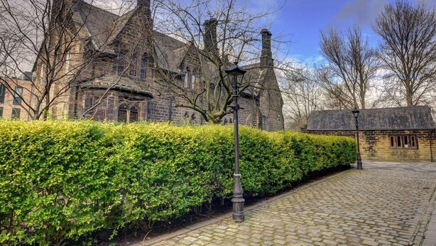 A historic stone building with multiple chimneys, a cobblestone pathway, and a well-maintained hedge. - Homebird Property - The Vicarage (Leeds)