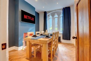 Un área de comedor con una mesa de madera, sillas, un televisor mostrando 'NETFLIX' y una ventana con cortinas.