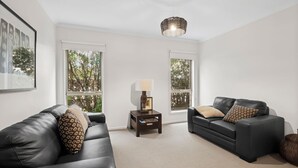 Una sala de estar moderna con dos sofás de cuero negro, una mesita de madera con una lámpara y grandes ventanales con vistas a árboles.