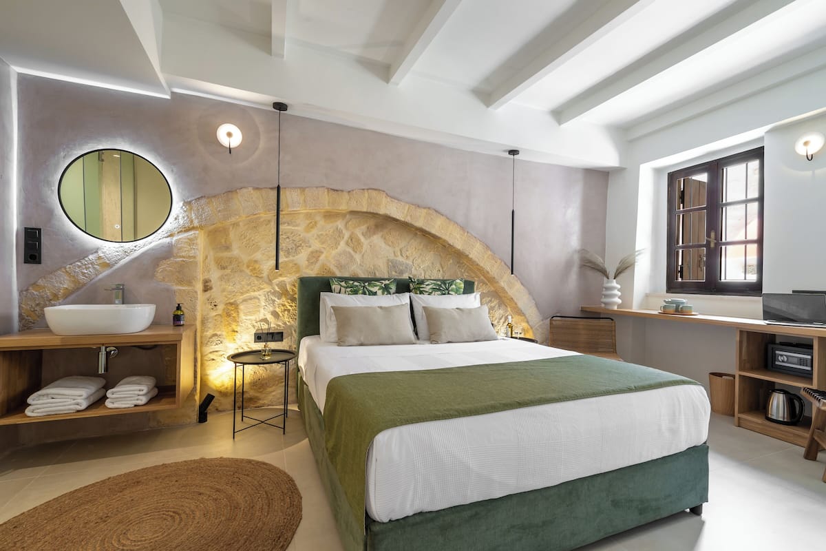 A modern bedroom with a stone arch, a large bed, a round mirror, a wooden vanity, and a desk with a computer.