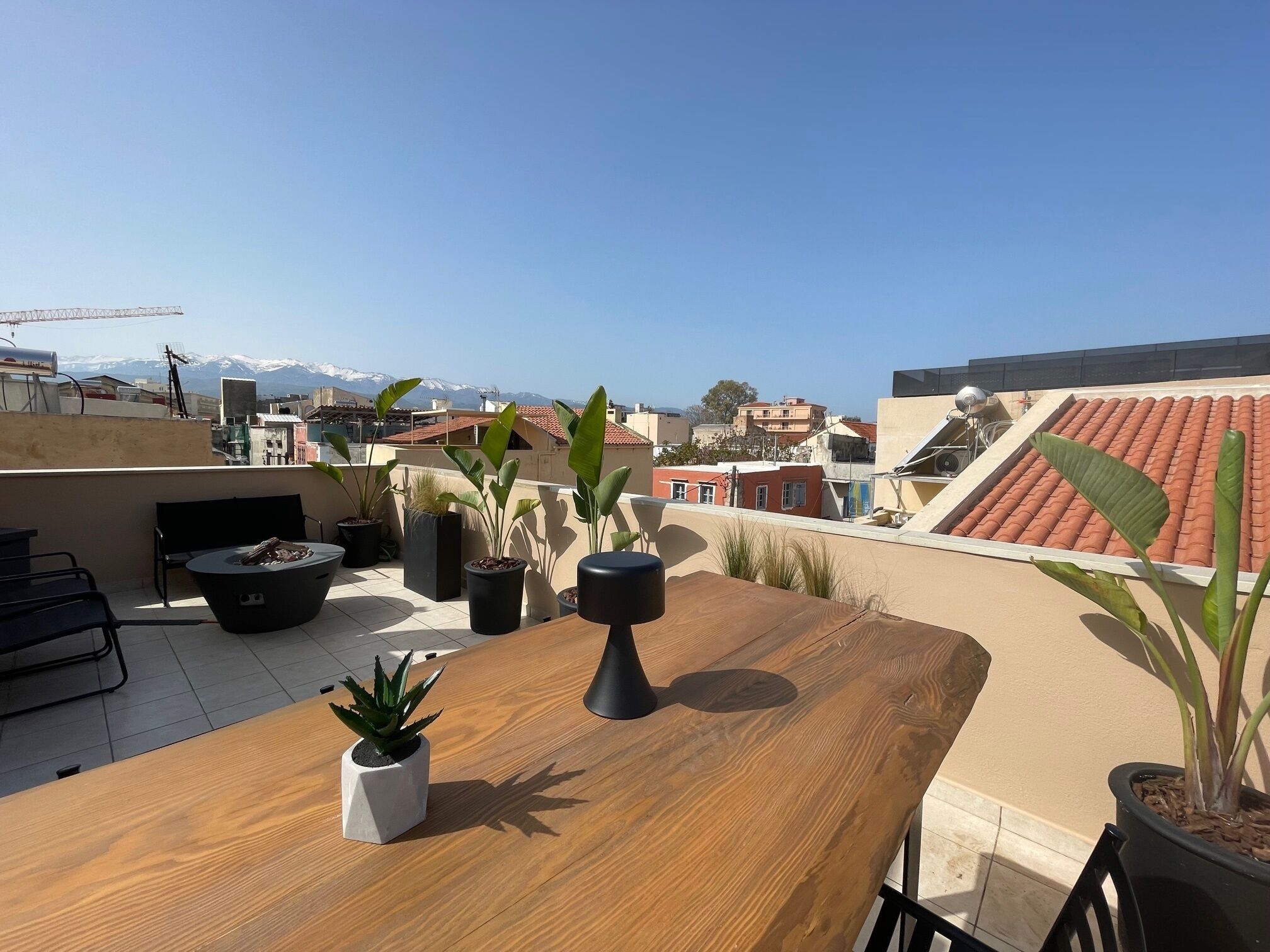 A rooftop terrace with a wooden table, potted plants, and a view of buildings and mountains.