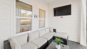A covered patio area with a sectional sofa, a small table with a potted plant, and a mounted TV on the wall.