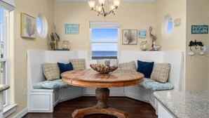 A corner seating area with a round wooden table, white cushioned benches, and a view of the ocean through a window.