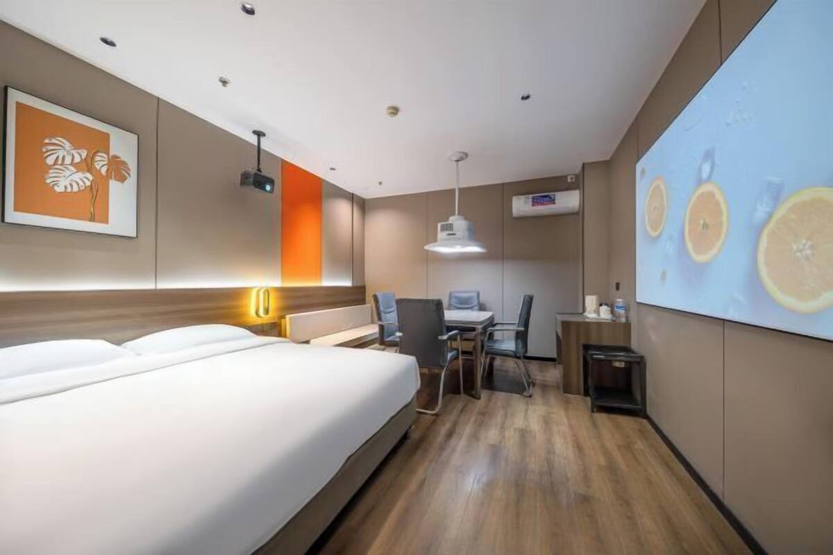 Une chambre d’hôtel équipée d’un lit, d’un bureau, de chaises et d’un écran de projection sur lequel sont affichés des fruits.