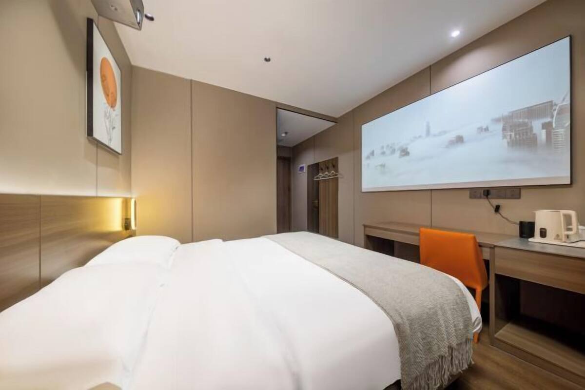 Une chambre d’hôtel moderne, équipée d’un lit, d’un bureau, d’une chaise, d’un grand écran et d’un tableau encadré au mur.
