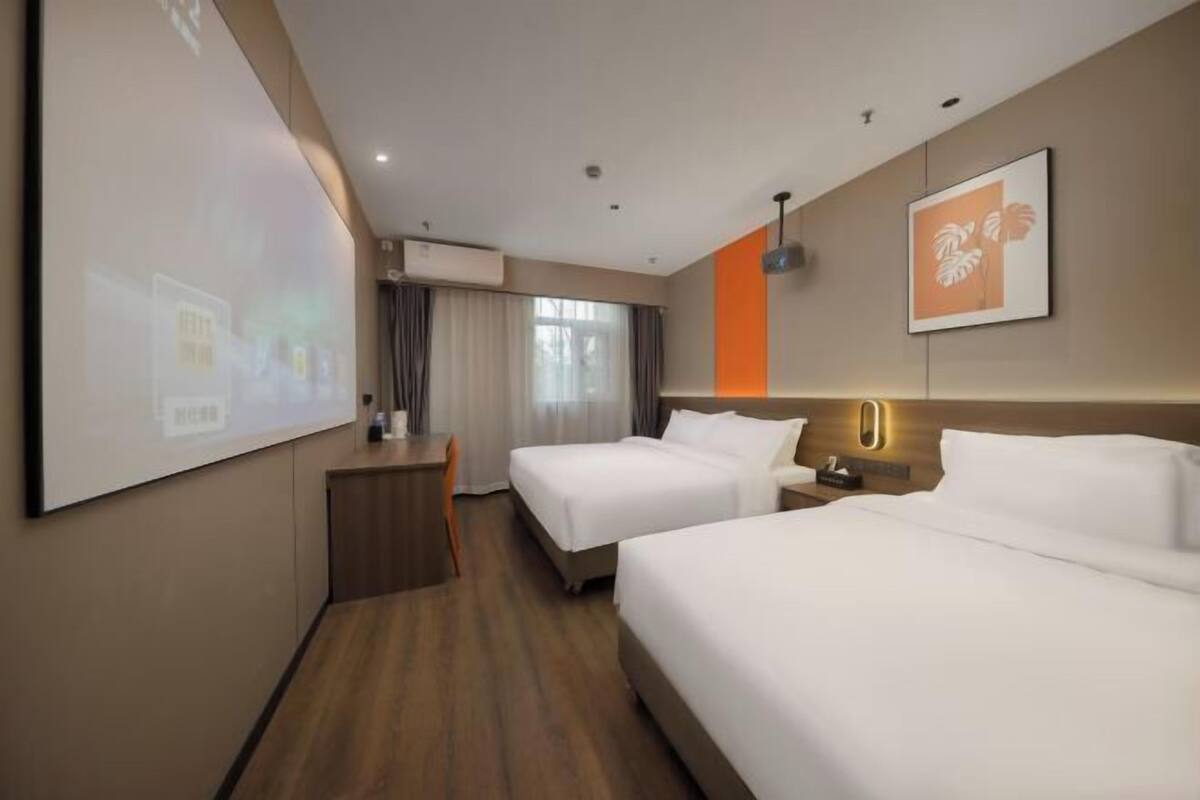 Une chambre d’hôtel avec deux lits, un bureau, une chaise, un écran de projection et un tableau encadré au mur.