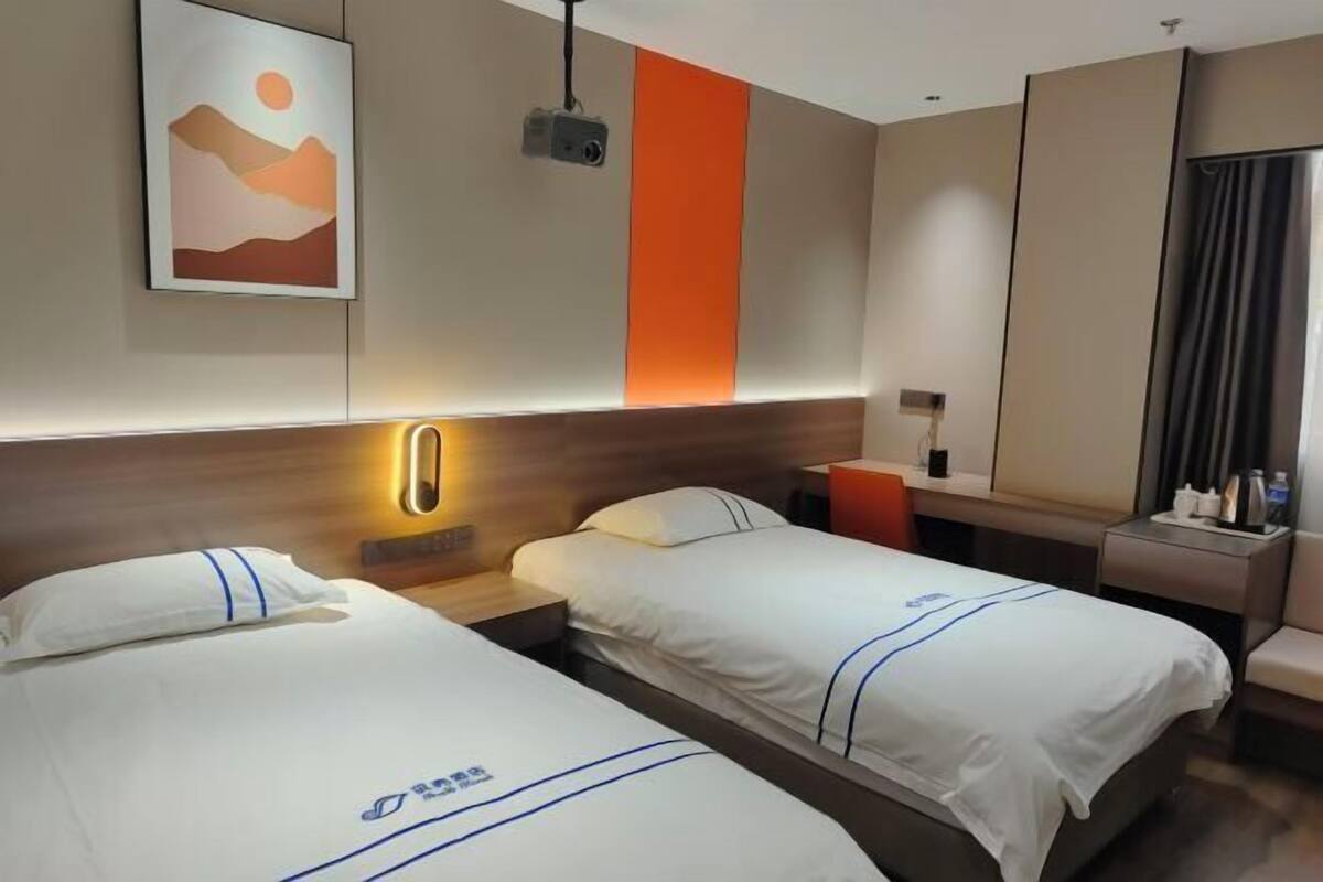 Une chambre d’hôtel avec deux lits, une table de chevet, un bureau, une chaise, un miroir et un projecteur fixé au mur.