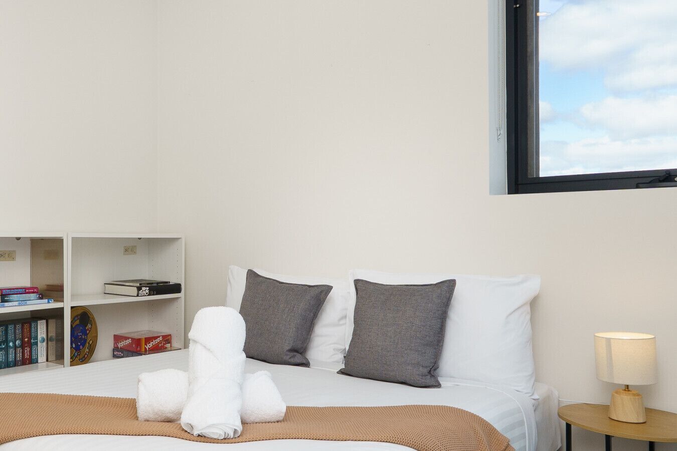 A neatly made bed with white and gray pillows, a rolled white towel, and a tan throw blanket. There is a small wooden nightstand with a lamp to the right. A bookshelf with books and a DVD case is visible to the left.