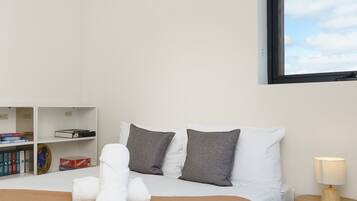 A neatly made bed with white and gray pillows, a rolled white towel, and a tan throw blanket. There is a small wooden nightstand with a lamp to the right. A bookshelf with books and a DVD case is visible to the left.
