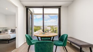 Een moderne kamer met een ronde tafel en vier groene stoelen, een bankje, en een uitzicht op gebouwen door grote ramen.
