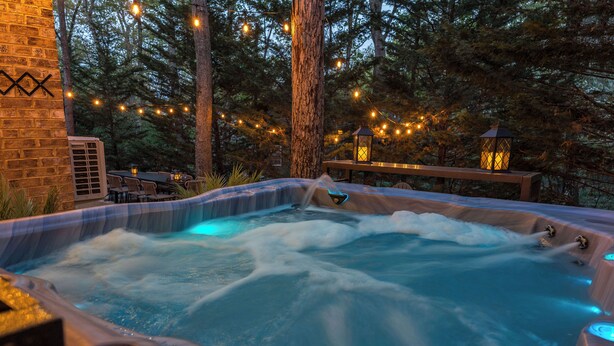 A hot tub surrounded by trees with string lights, a wooden deck, and a brick wall.