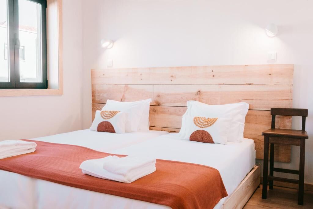 A hotel room with a wooden headboard, two beds with white bedding and brown pillows, a wooden nightstand, and a window with blinds.