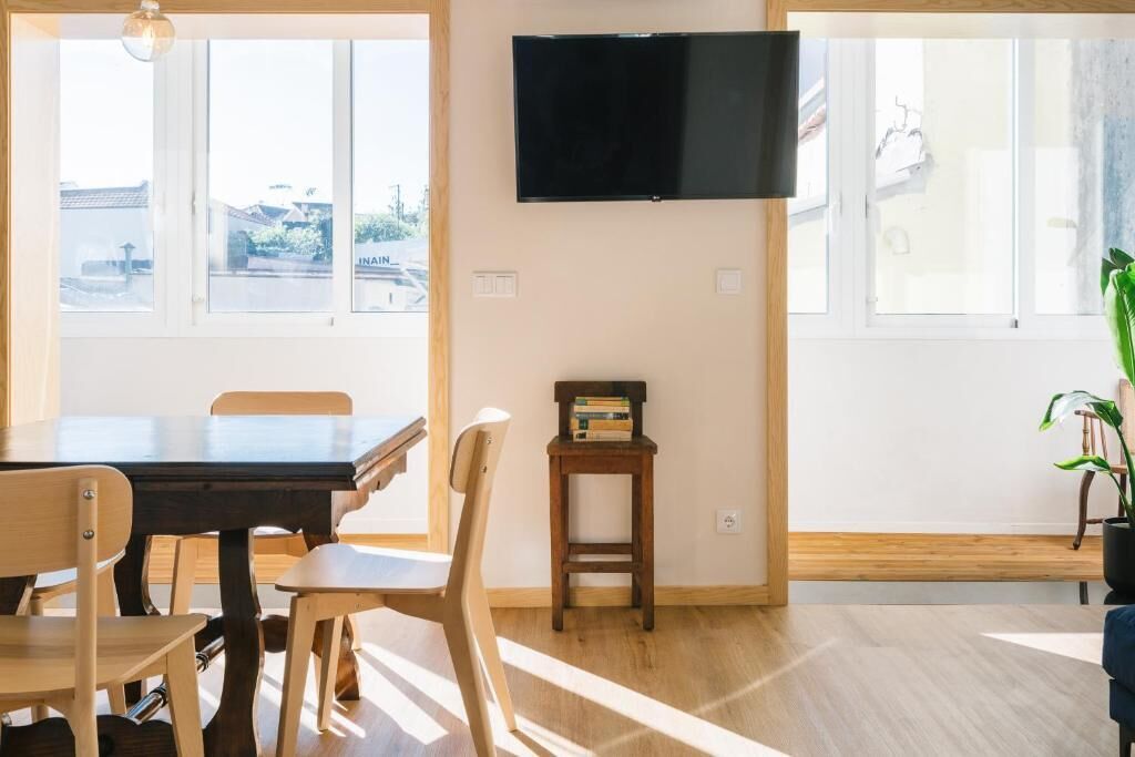 A room with a dining table, chairs, a small wooden stool, a television mounted on the wall, and a potted plant.