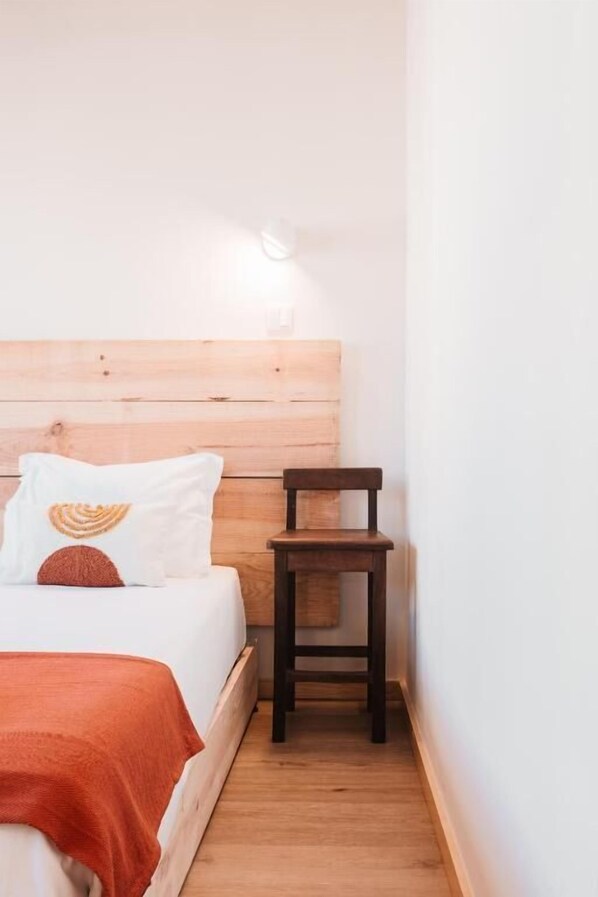 Un dormitorio con cabecera de madera, cama con ropa blanca y una manta naranja, y una pequeña silla de madera.