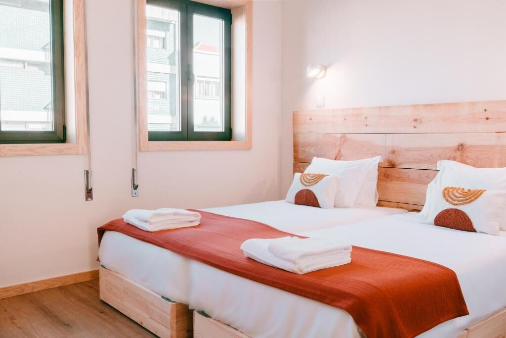 A double bed with a wooden headboard, white bedding, and a red throw blanket. There are two windows with wooden frames and a view of buildings outside.