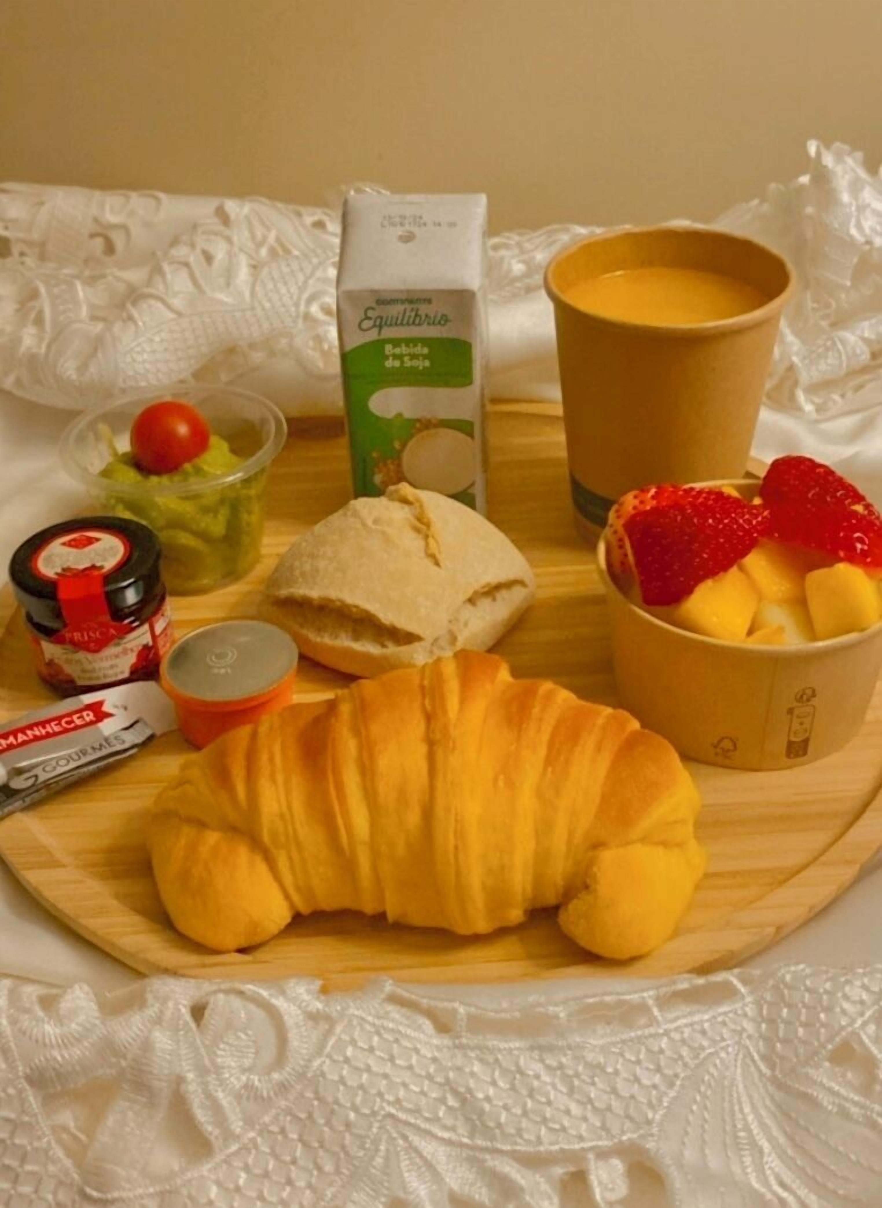 Bandeja de desayuno con un croissant, una taza de café, un tazón de fruta, un recipiente de yogur, un frasco pequeño de mermelada y un paquete de ManHecher.