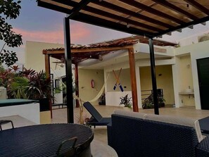 Relax in a shaded patio area with lounge chairs, a hammock, and a small table, surrounded by lush potted plants.