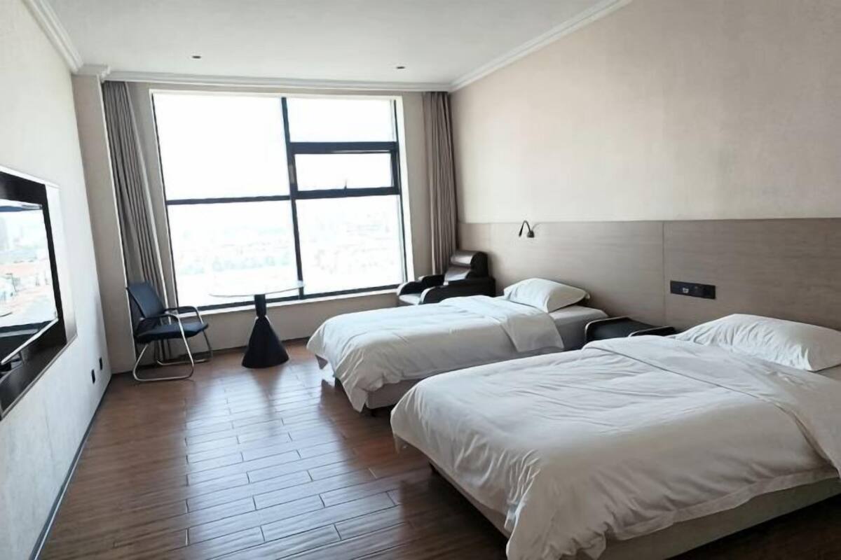 A hotel room with two beds, a desk, a chair, a window with curtains, and a view of the city.