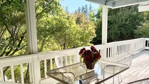 An outdoor patio with a glass table and white wicker chairs, surrounded by a white railing and lush greenery.
