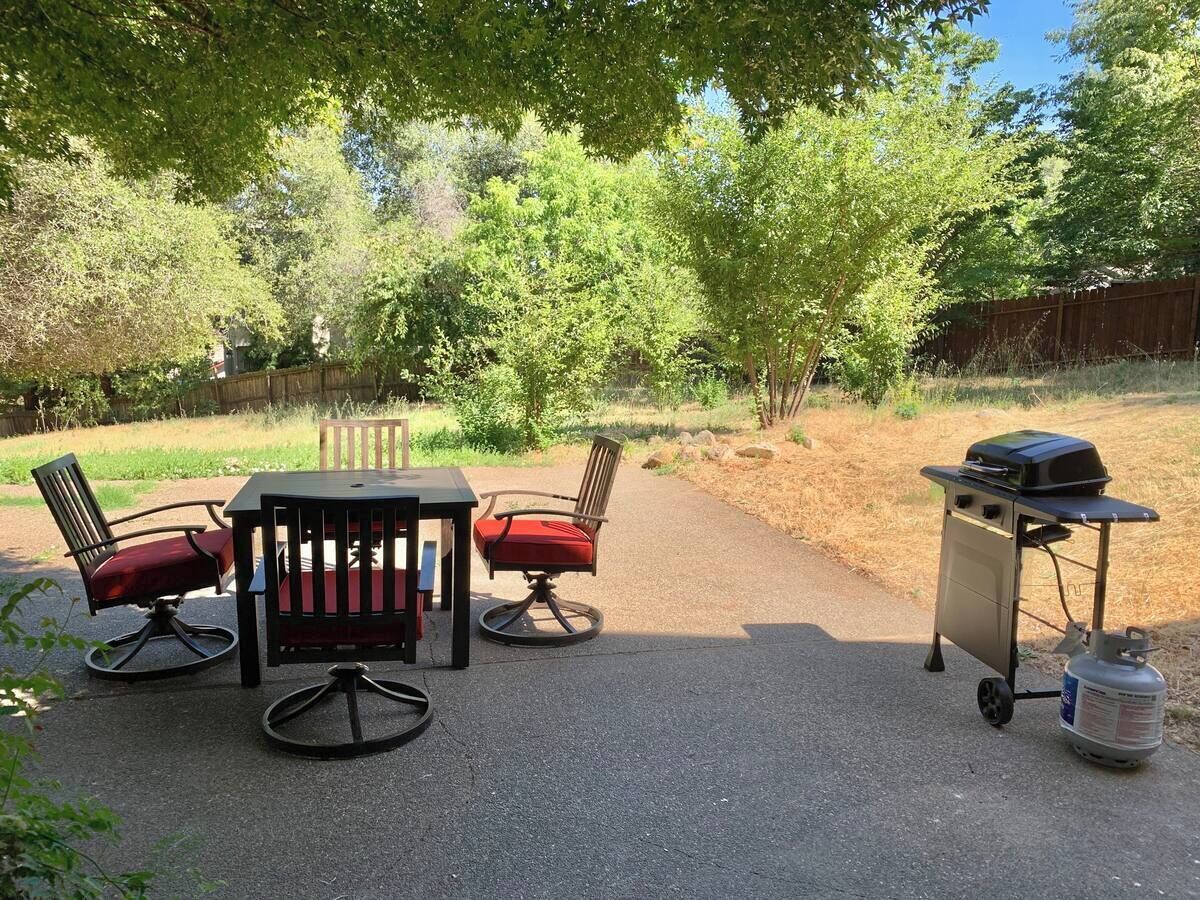 En udendørs terrasse med et bord, to stole, en grillvogn og en propangasflaske.