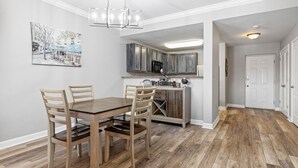 A dining area with a wooden table and chairs, a kitchen with grey cabinets, and a hallway with a white door. - Eagles Landing 1BR Heaven (Pigeon Forge)