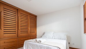 A bedroom with a bed, white bedding, a wooden wardrobe, and a white wall.