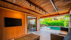 A modern living room with a flat-screen TV, wicker furniture, and a view of a swimming pool. - Bz01 Casa Piscina Priv 150m Da Praia  (Buzios)