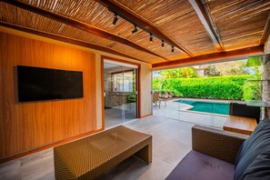 A modern living room with a flat-screen TV, wicker furniture, and a view of a swimming pool. - Bz01 Casa Piscina Priv 150m Da Praia  (Buzios)