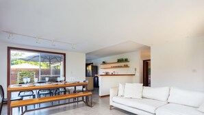 A modern living room with a white sofa, wooden dining table, and chairs. There is a kitchen area in the background with a refrigerator and shelves. - Bz01 Casa Piscina Priv 150m Da Praia  (Buzios)
