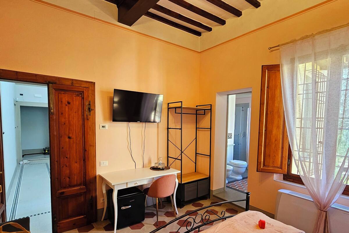Une chambre à coucher avec un lit, un bureau et une télévision. Il y a une fenêtre avec des rideaux et une vue sur l’extérieur.