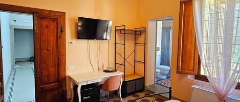 A bedroom with a bed, a desk, and a TV. There is a window with curtains and a view of the outside.
