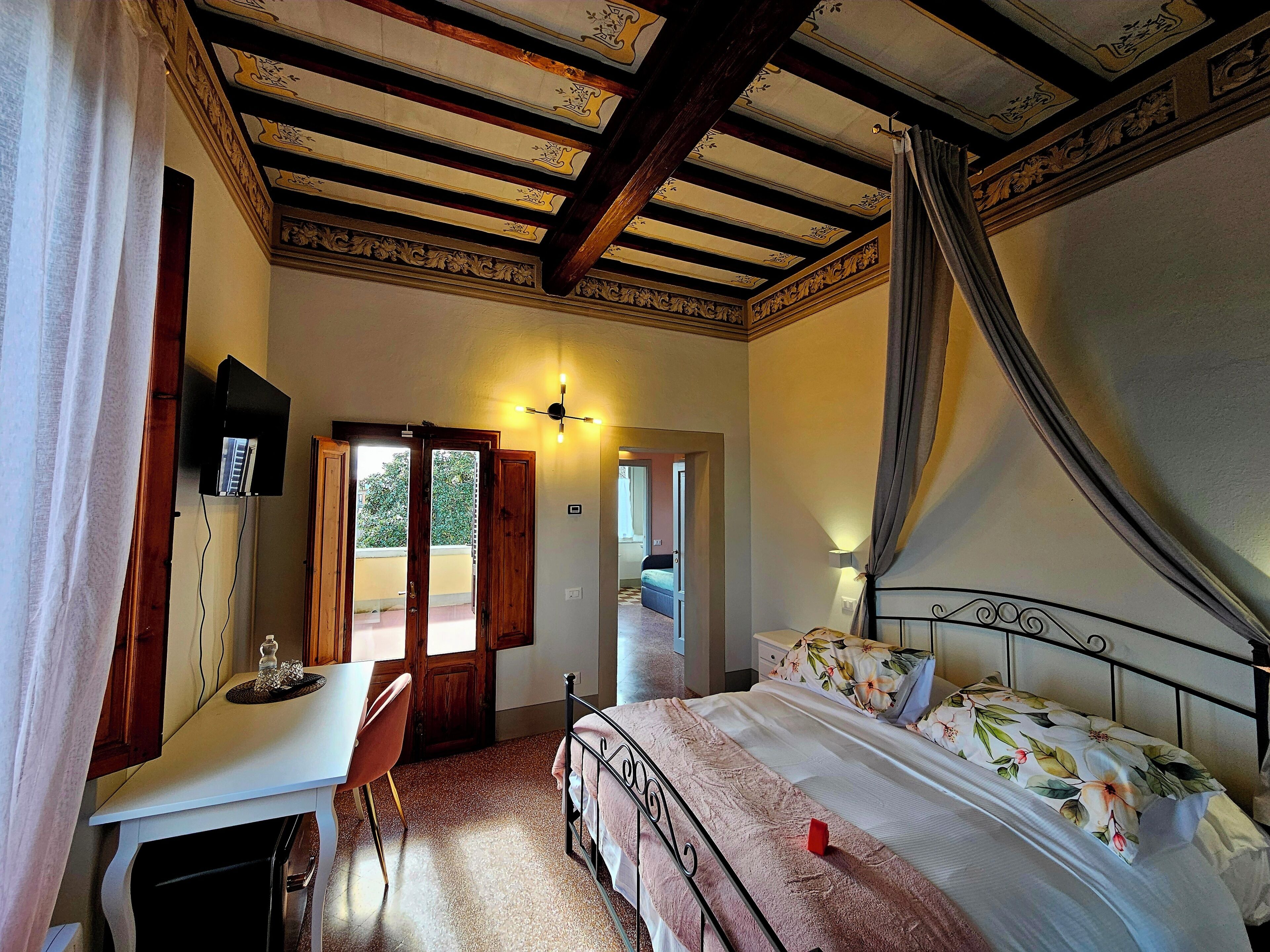 A bedroom with a bed, a desk, a chair, a television, and a ceiling with decorative beams.