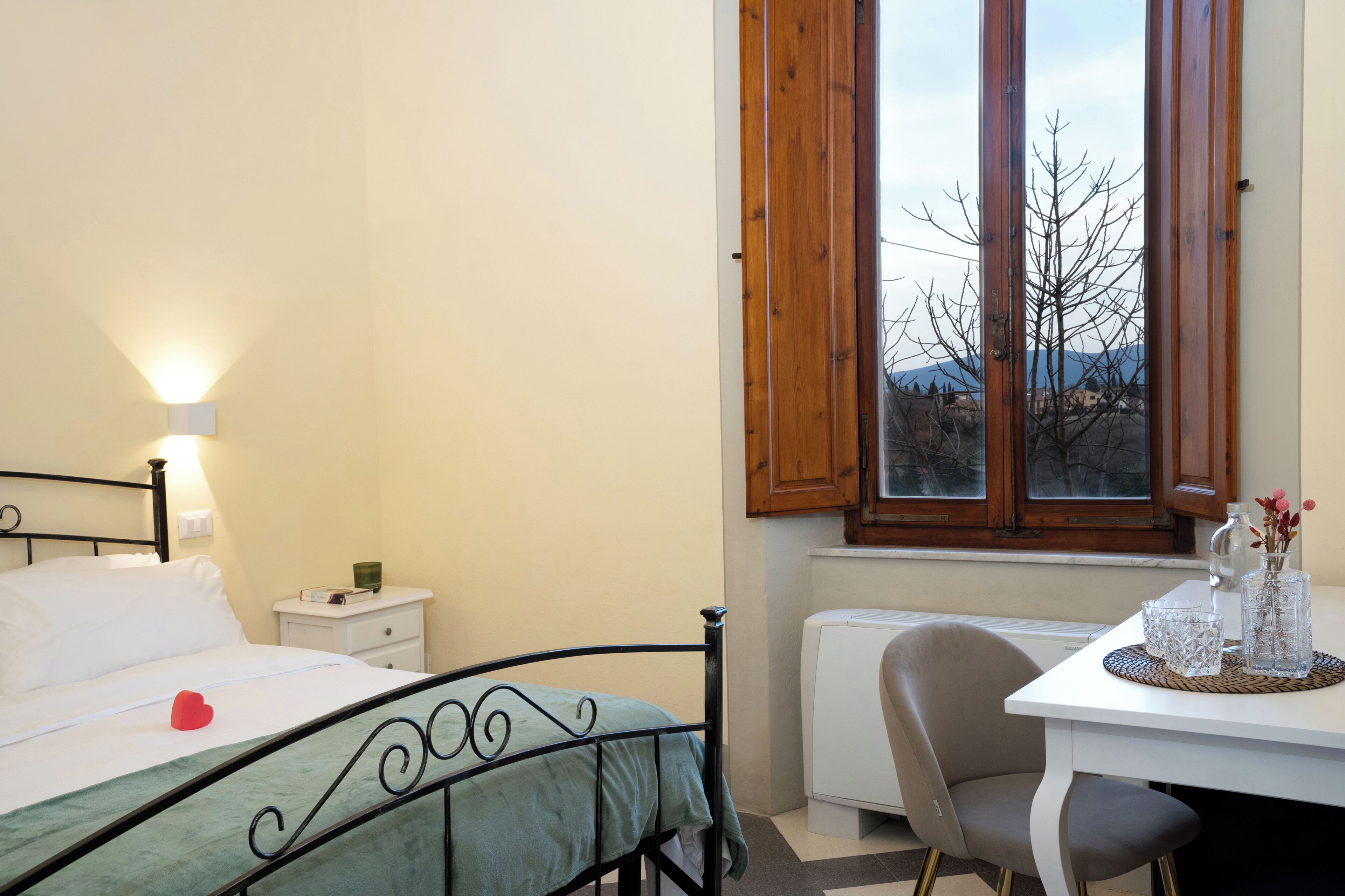 Una camera da letto con un letto, un comodino, una lampada, una finestra, un termosifone e una scrivania con una sedia.