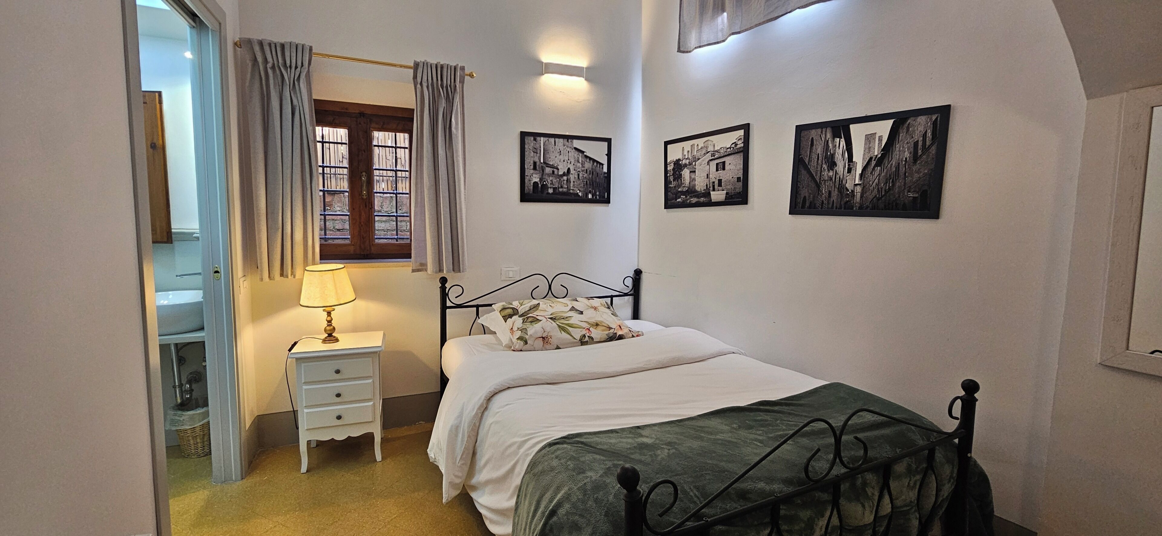 A bedroom with a bed, bedside table, lamp, curtains, and three black-and-white photographs on the wall.