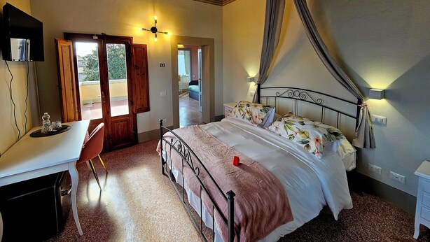 A bedroom with a bed, a desk, and a chair. There is a door leading to another room and a window with curtains.