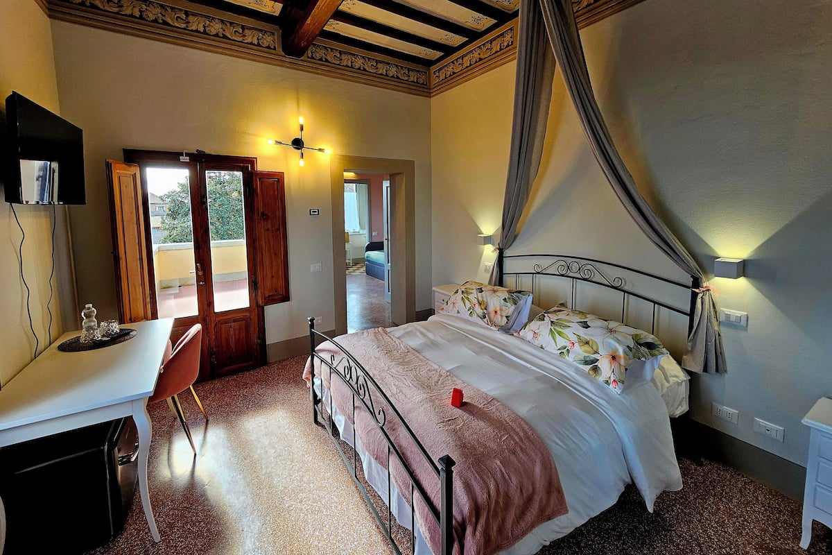 Une chambre à coucher avec un lit, un bureau et une chaise. Il y a une porte qui donne sur une autre pièce et une fenêtre avec des rideaux.