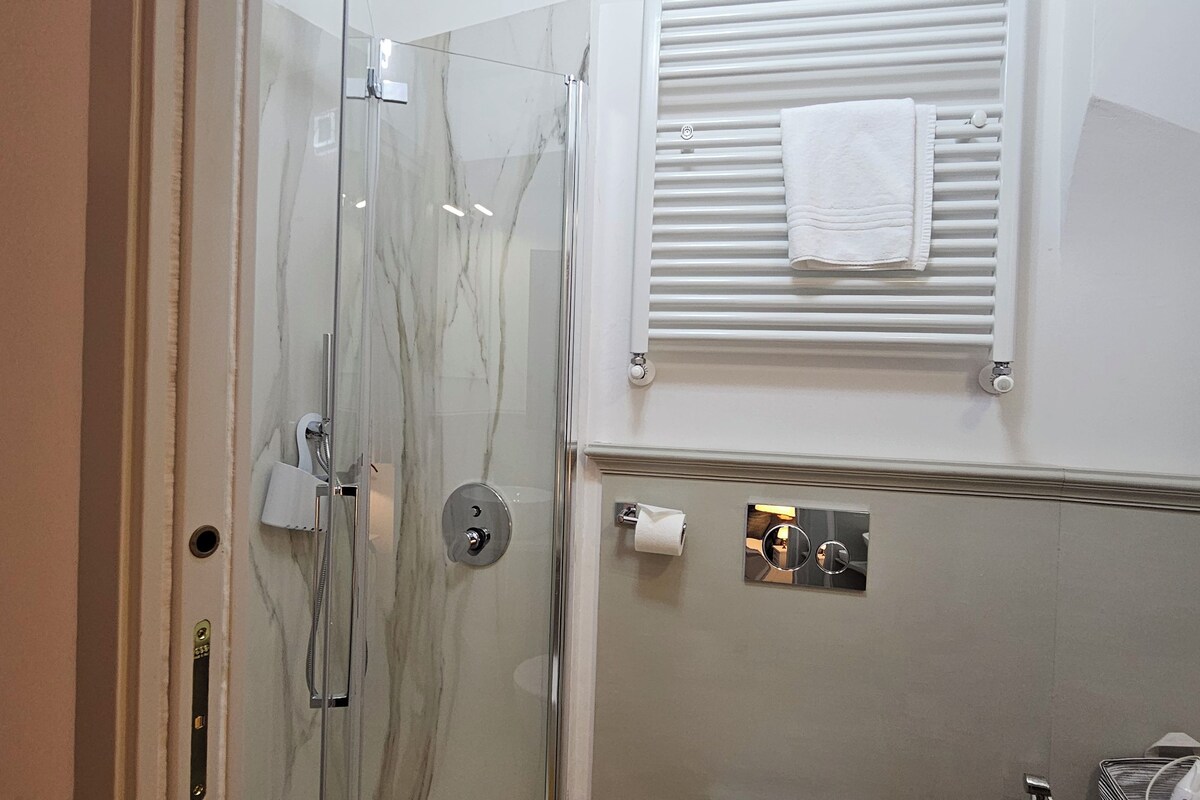 Ein Badezimmer mit einer gläsernen Duschkabine, einem Handtuchhalter, einem Toilettenpapierrollenhalter und einem kleinen Fenster mit Jalousien.