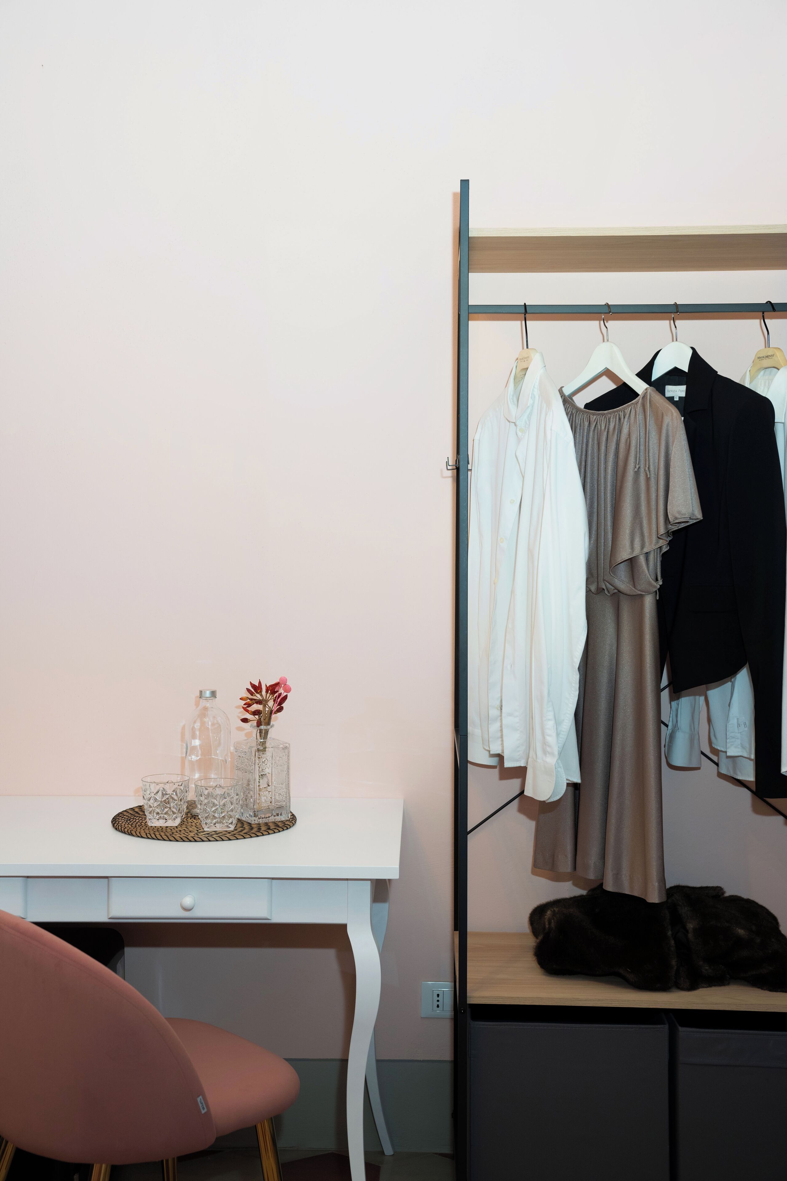 A room with a white desk, a pink chair, a clothing rack with various garments, and a small table with decorative items.