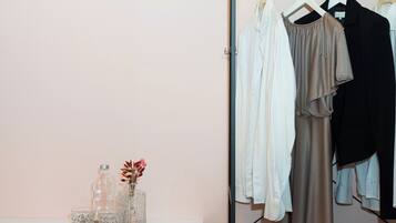 A room with a white desk, a pink chair, a clothing rack with various garments, and a small table with decorative items.