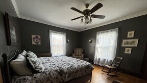 Une chambre à coucher avec un lit, une chaise, un ventilateur de plafond et une fenêtre avec des rideaux.