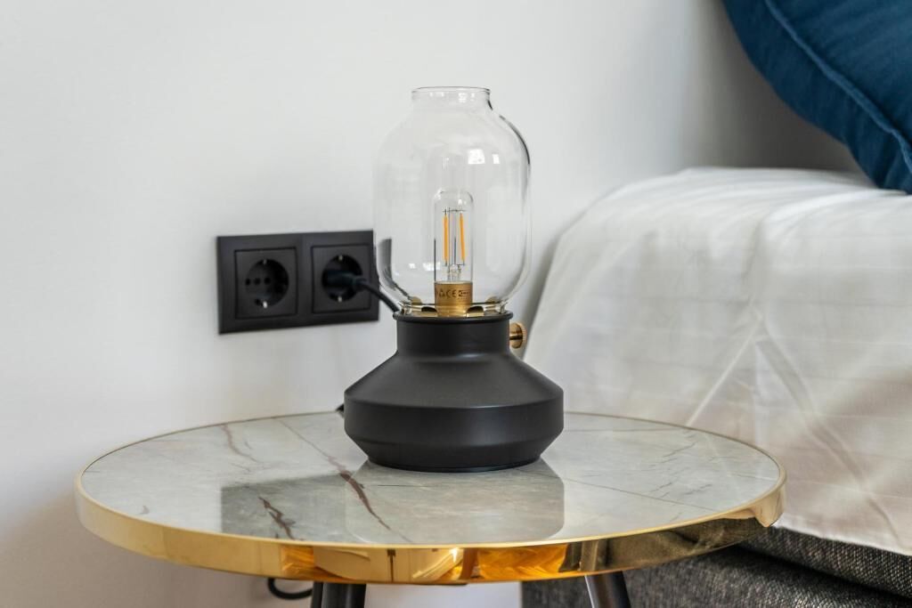 En moderne lampe med klart glasskabinet på et marmorbord, en stikkontakt og en seng med hvidt sengetøj i baggrunden.